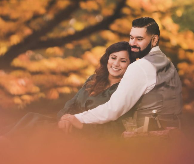 The engaged couple standing together on a manicured lawn, with a large, historic red-brick building and manicured gardens in the background.