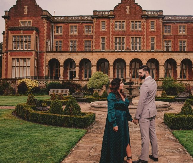 The couple standing on an ornate balcony overlooking a formal garden with a large historic building in the background.