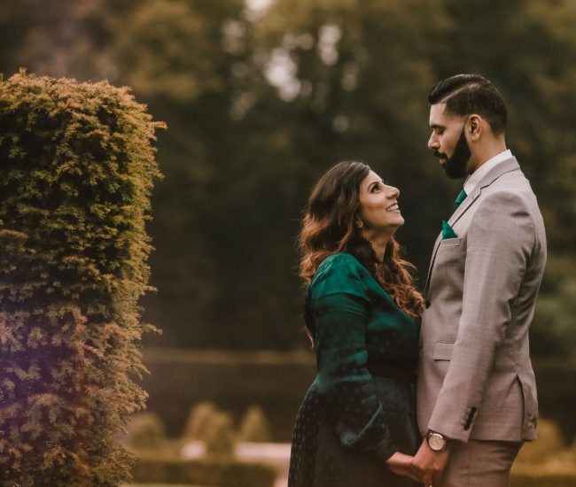 A pre-wedding photo of a couple standing side by side, looking into a green, lush garden setting.
