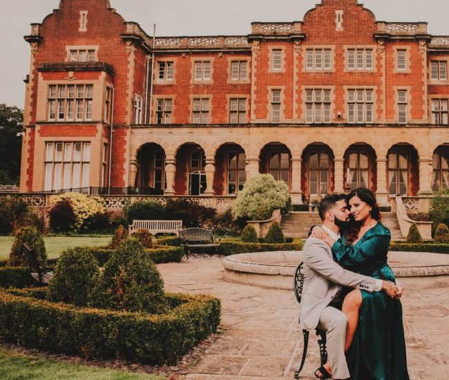 formal pre-wedding photo of a couple sitting at a small table outdoors, with the groom looking at the bride, who is smiling.