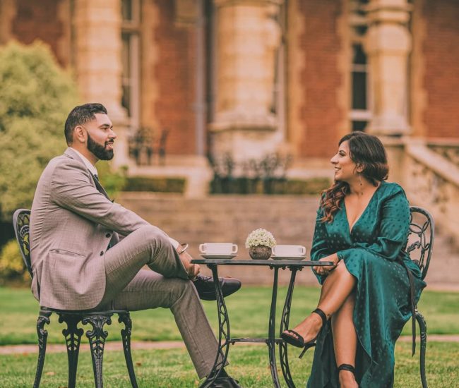 A couple sitting on a garden bench with a historic red-brick building and manicured hedges in the background, captured during their pre-wedding photoshoot.