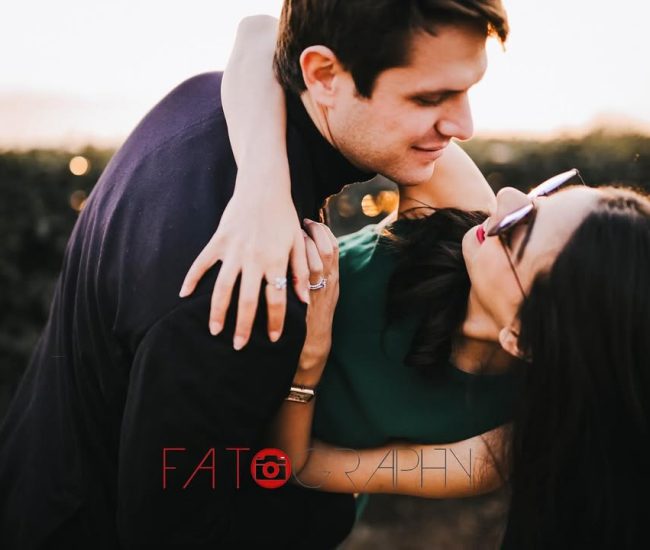 A close-up portrait of the couple embracing in a natural field setting, with a warm backlight and a blurred background.