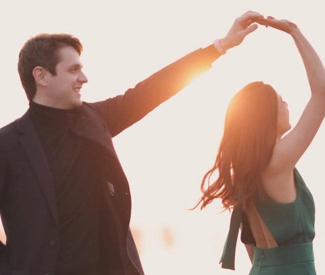 The couple standing side-by-side with the bride's arm outstretched, as if in a playful dance, in a field setting.