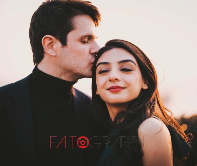 The groom standing behind his bride, holding her close while she looks at the camera, with a warm, romantic backdrop.