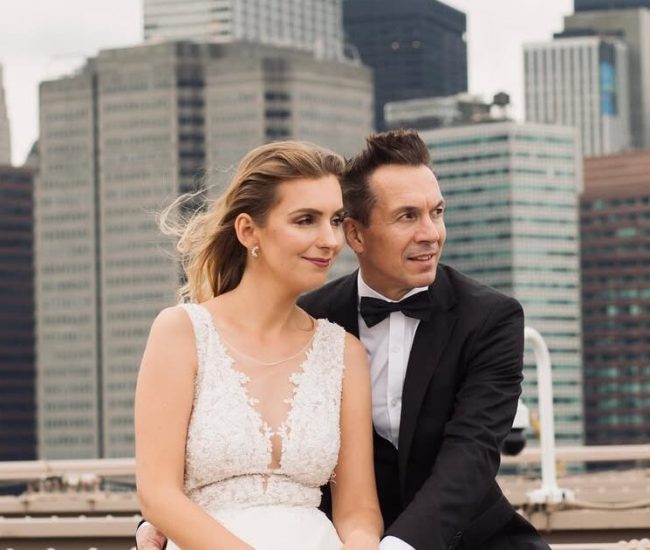 Groom leaning on bridge railing with city skyline in background during Fatography pre wedding shoot.