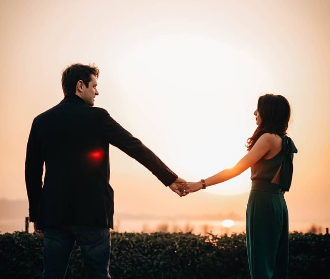 An engaged couple walking hand-in-hand in silhouette against a warm, hazy sunset sky during their romantic pre-wedding photoshoot.