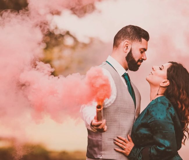 An engaged couple walking hand-in-hand past a tiered fountain in a formal garden, with an ornate building in the background.