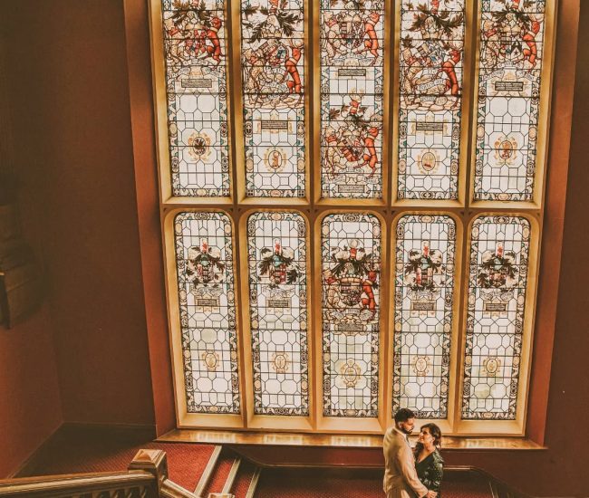 The bride and groom posing in front of a large ornate window with beautiful architectural details during an indoor portion of their pre-wedding photoshoot.