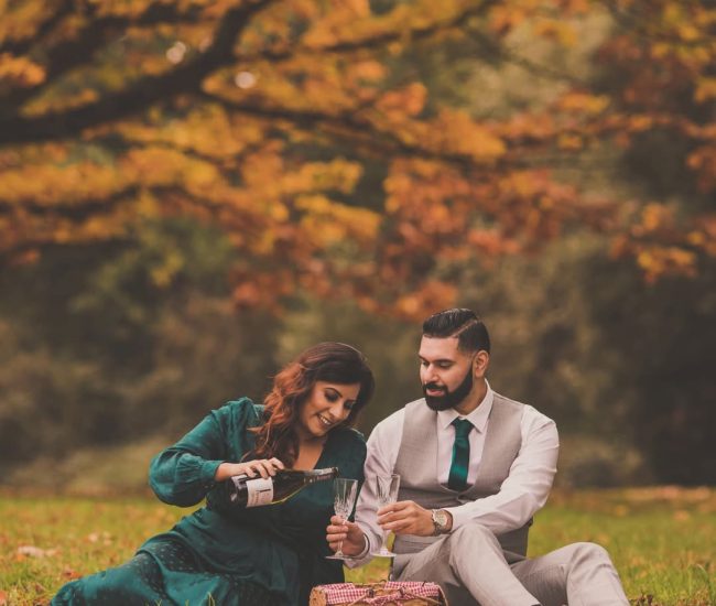 A pre-wedding photo of the groom wrapping his arms around his bride from behind while standing in a wooded area.